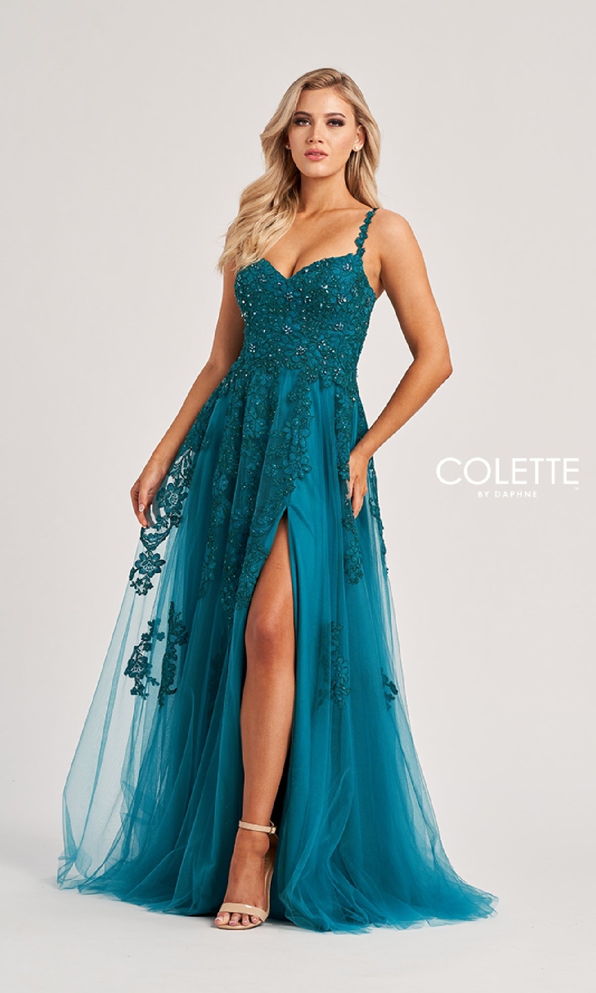 A woman wears the Colette CL8220 long prom dress featuring teal embroidery, lace detailing, thin straps, a sweetheart neckline, and a high front slit. The background is plain and light.